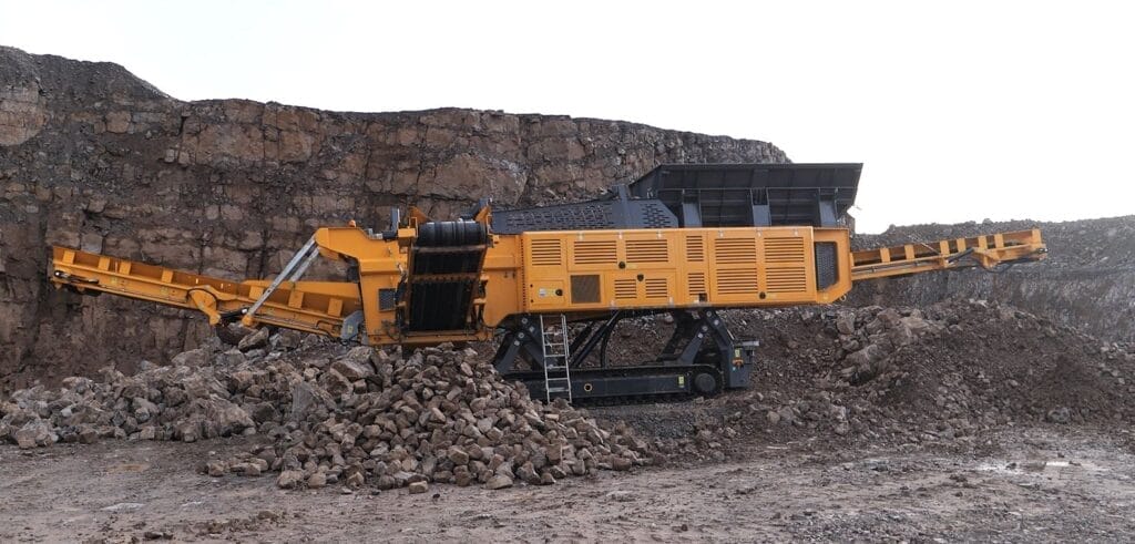 ranoworks A large yellow mobile rock crusher and screeners operate at a quarry site, surrounded by piles of broken rocks and stone walls in the background.
