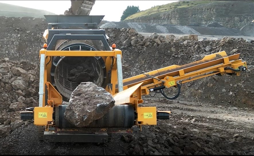 ranoworks A large rock is being fed into an industrial rock crusher at a Ranoworks quarry site, with screeners, a conveyor belt, and rough terrain in the background.